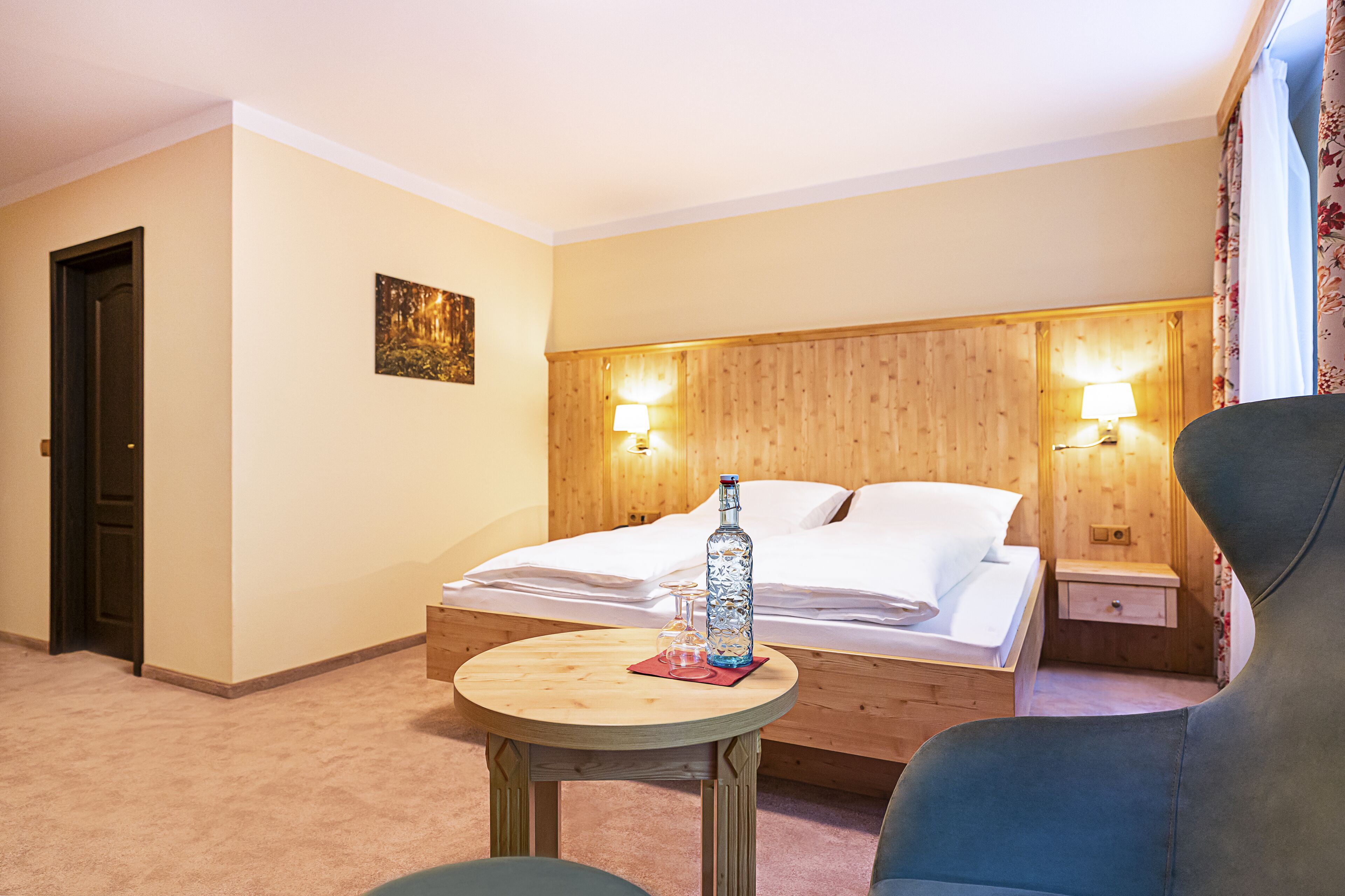 A hotel room with a bed, bedside lamps, a table with a bottle and glasses, and a chair.