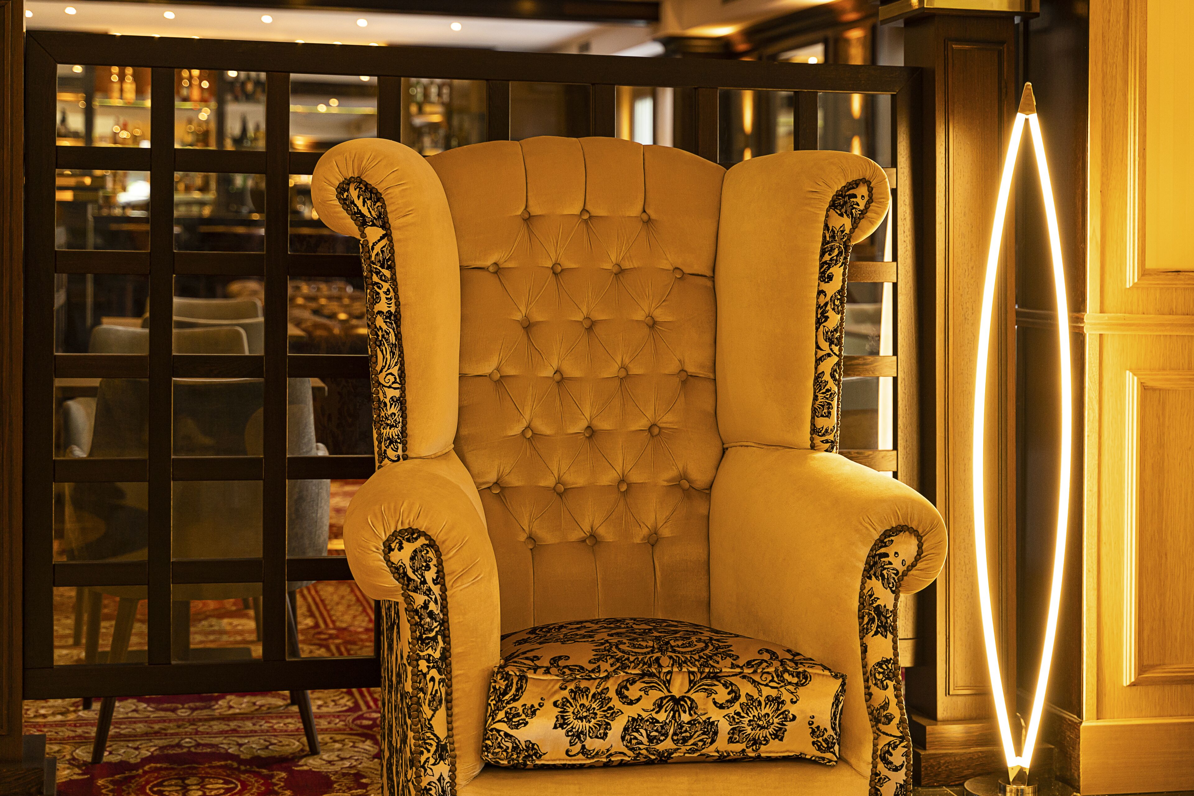 Ein vielfach verziertes Armchair mit einem gemusterten Kissen in einem wohlig beleuchteten Raum mit einem gemusterten Boden und einer Glasabtrennung.