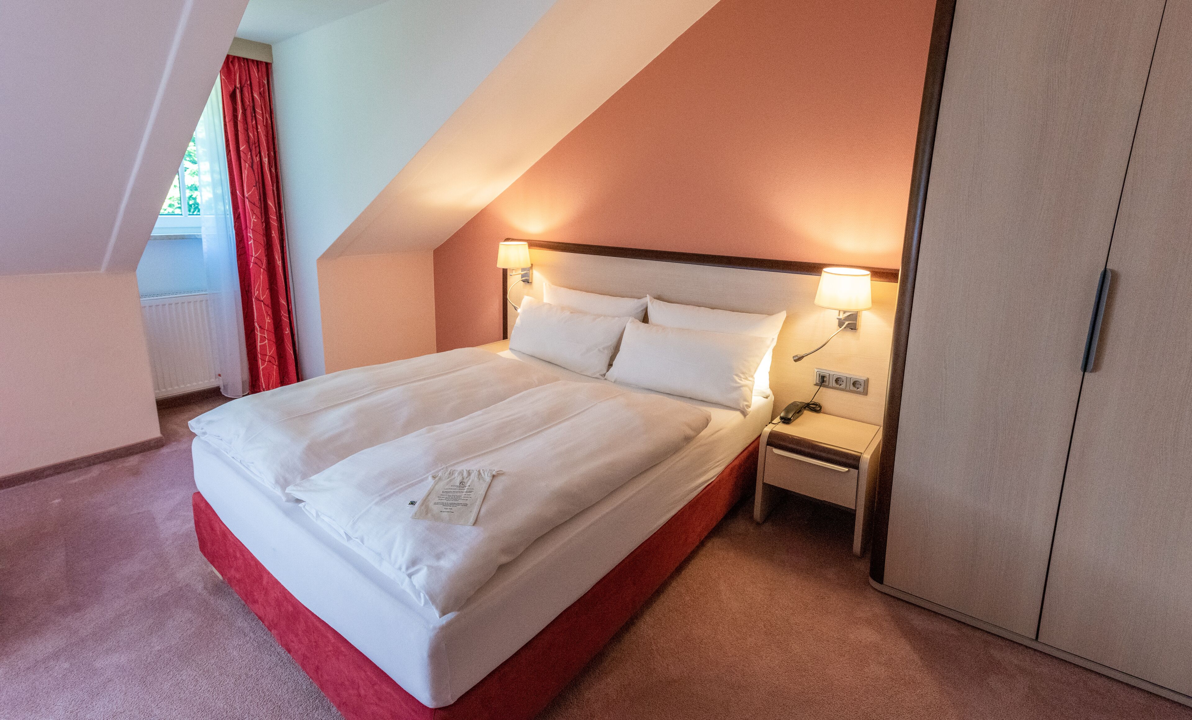 A neatly made bed with white linens in a hotel room with a sloped ceiling and a red accent wall.