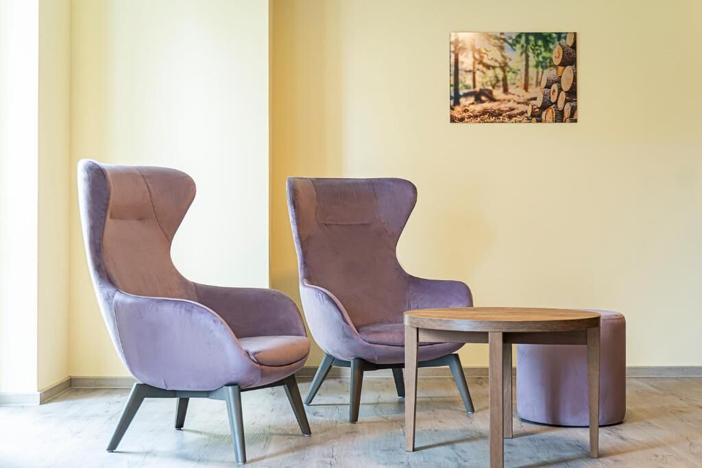 Two purple chairs and a small wooden table in a room with a light yellow wall and a painting of a forest.