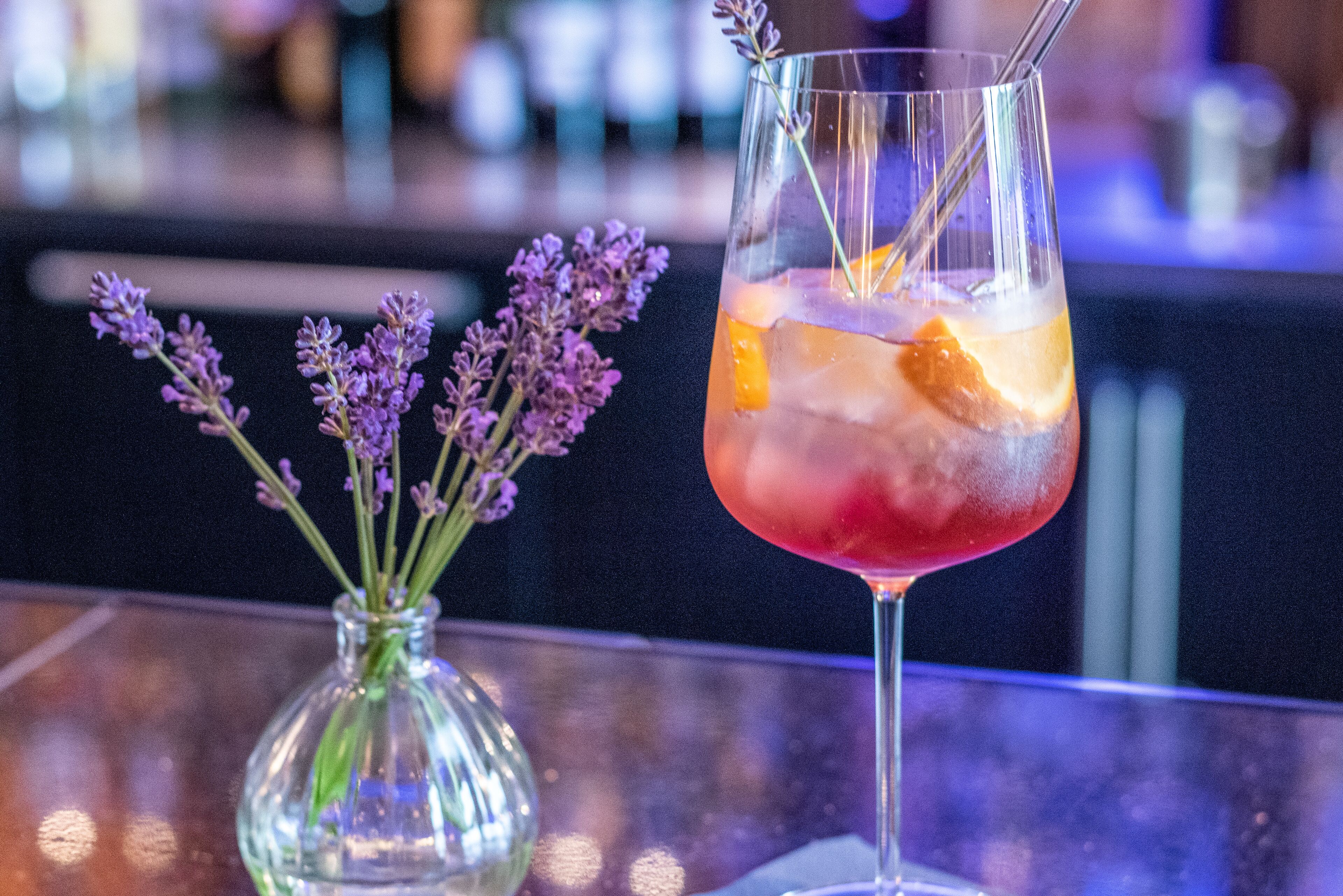 Een glas rode cocktail met ijs en een oranjeschijfje, samen met een vaas paarse bloemen op een bargedeelte.