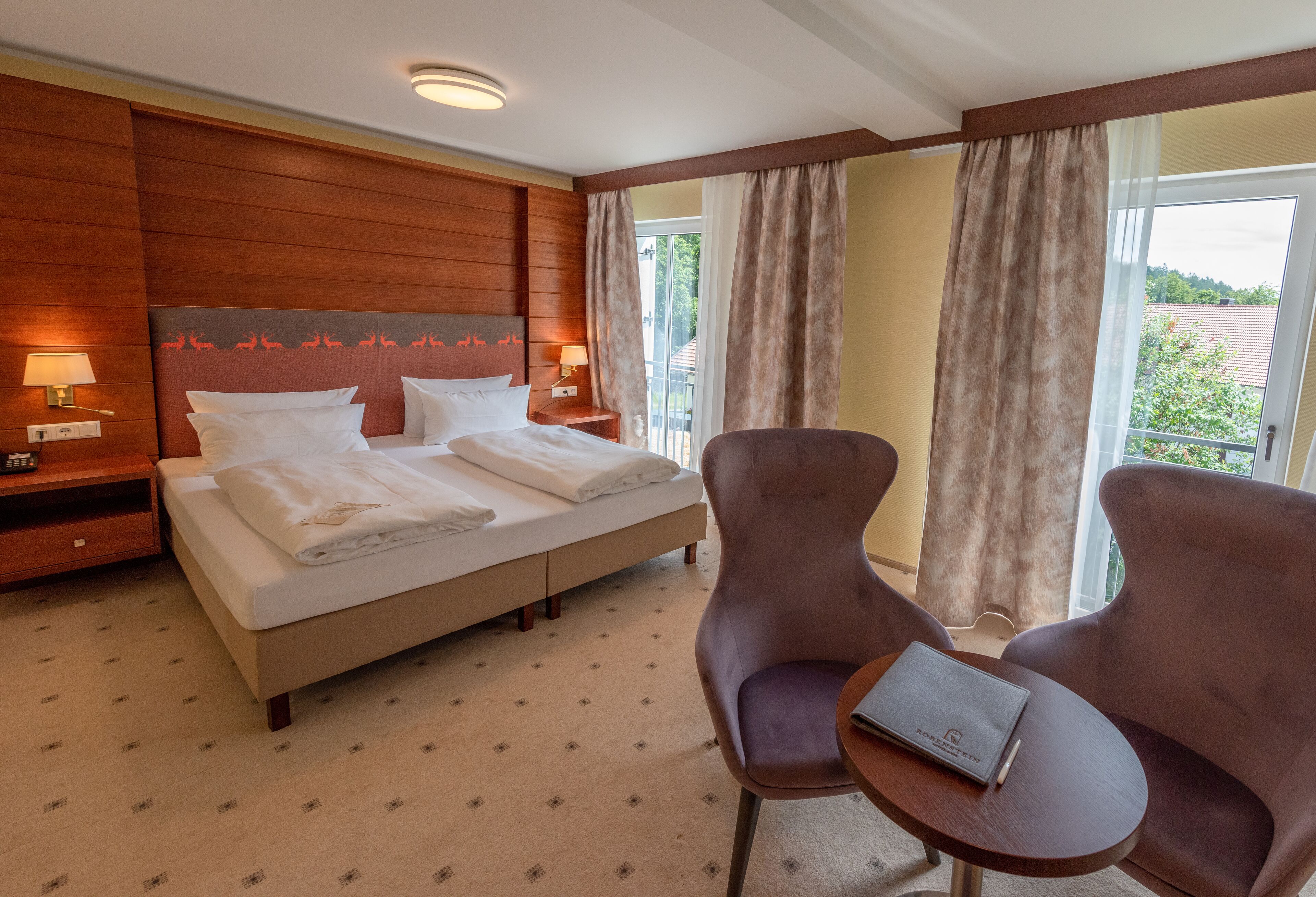 Una habitación de hotel con una cama grande, dos mesitas de noche con lámparas, una pequeña mesa redonda con un libro y vistas al exterior a través de un amplio ventanal.