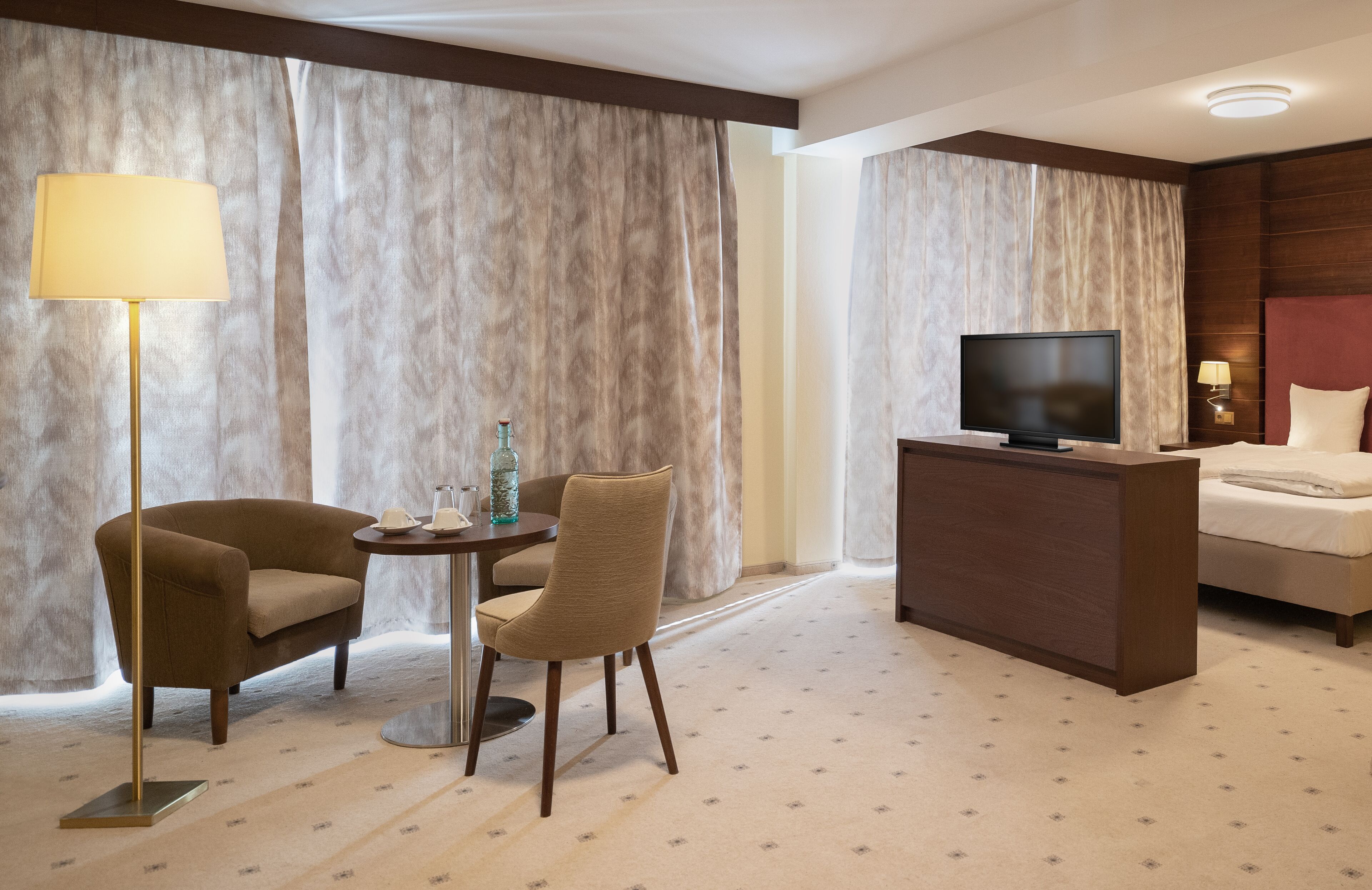 A hotel room with a bed, a desk with a television, a lamp, two chairs, and a small table with a bottle and glass.