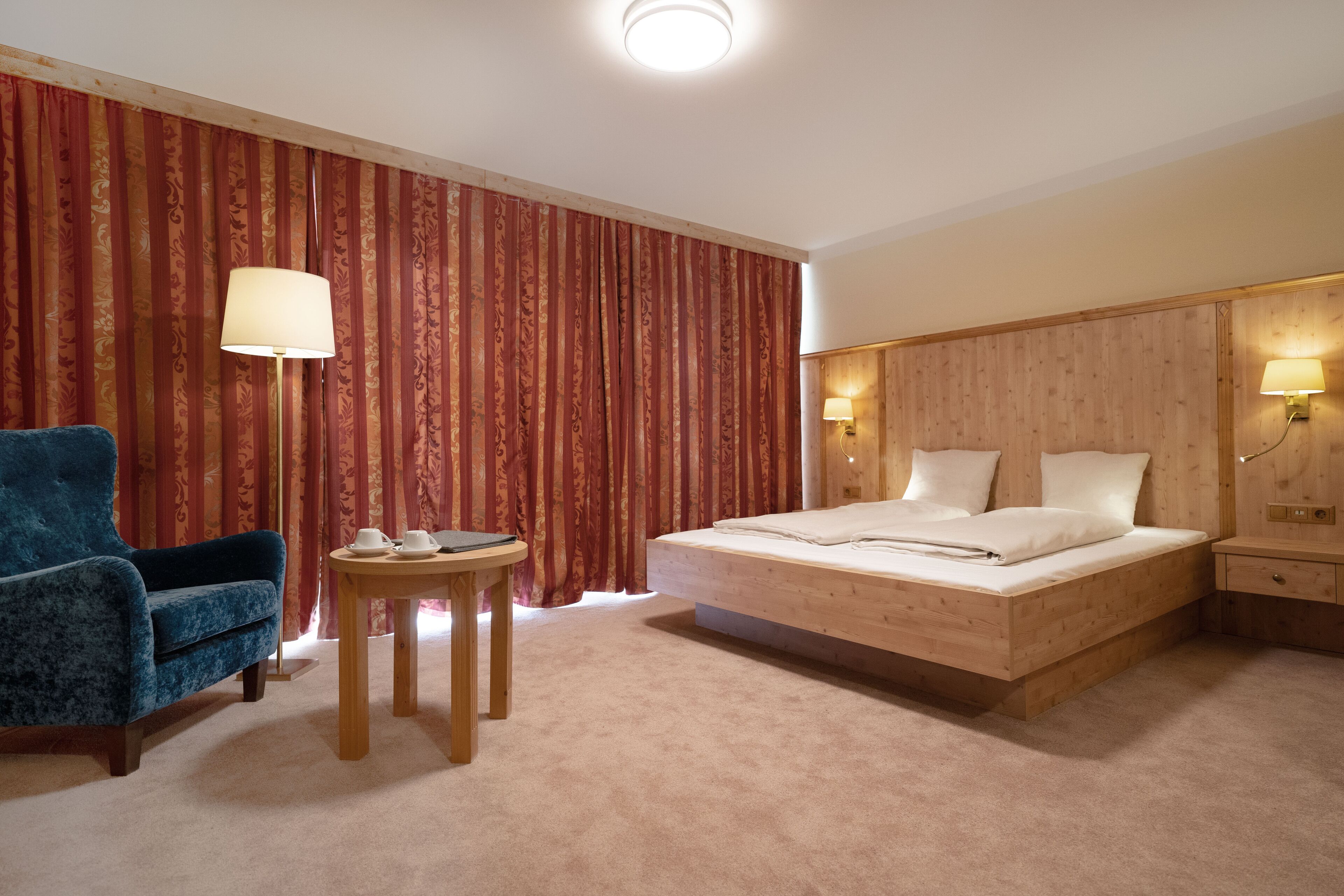Une chambre d’hôtel avec un lit, un fauteuil bleu, une petite table avec une lampe et des rideaux.