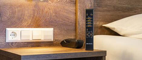 A wooden bedside table with a remote control, a wall socket, and a power outlet.