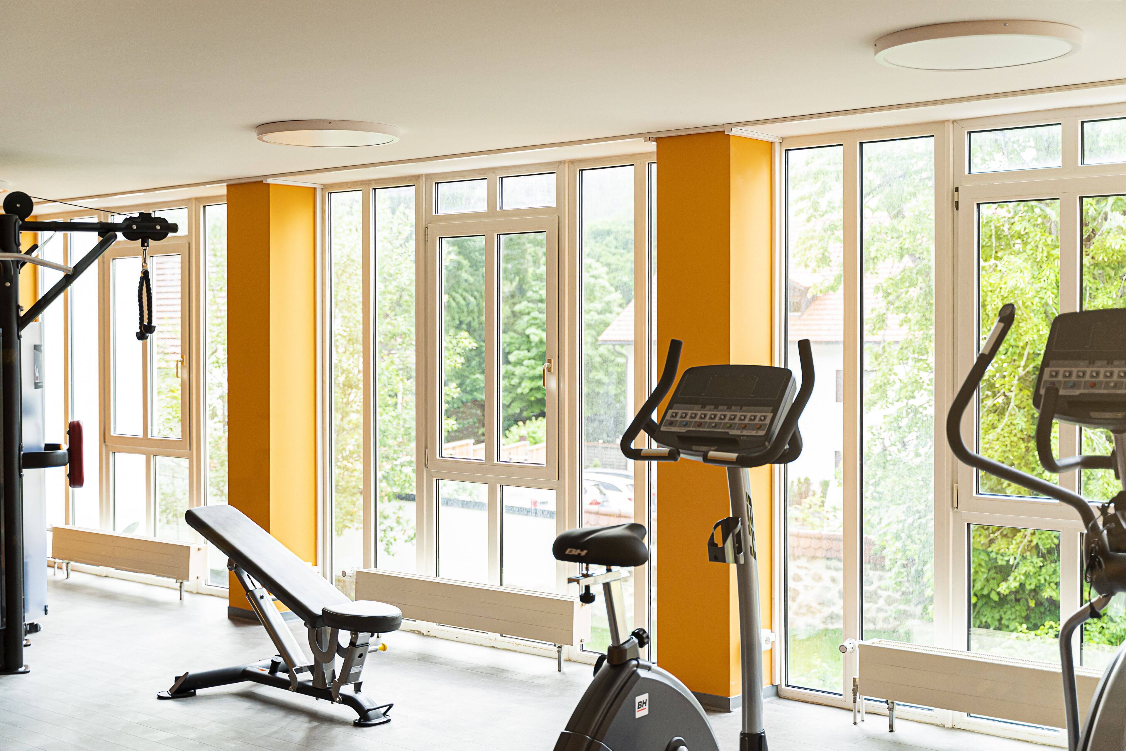 A gym with exercise equipment, large windows, and a bench.