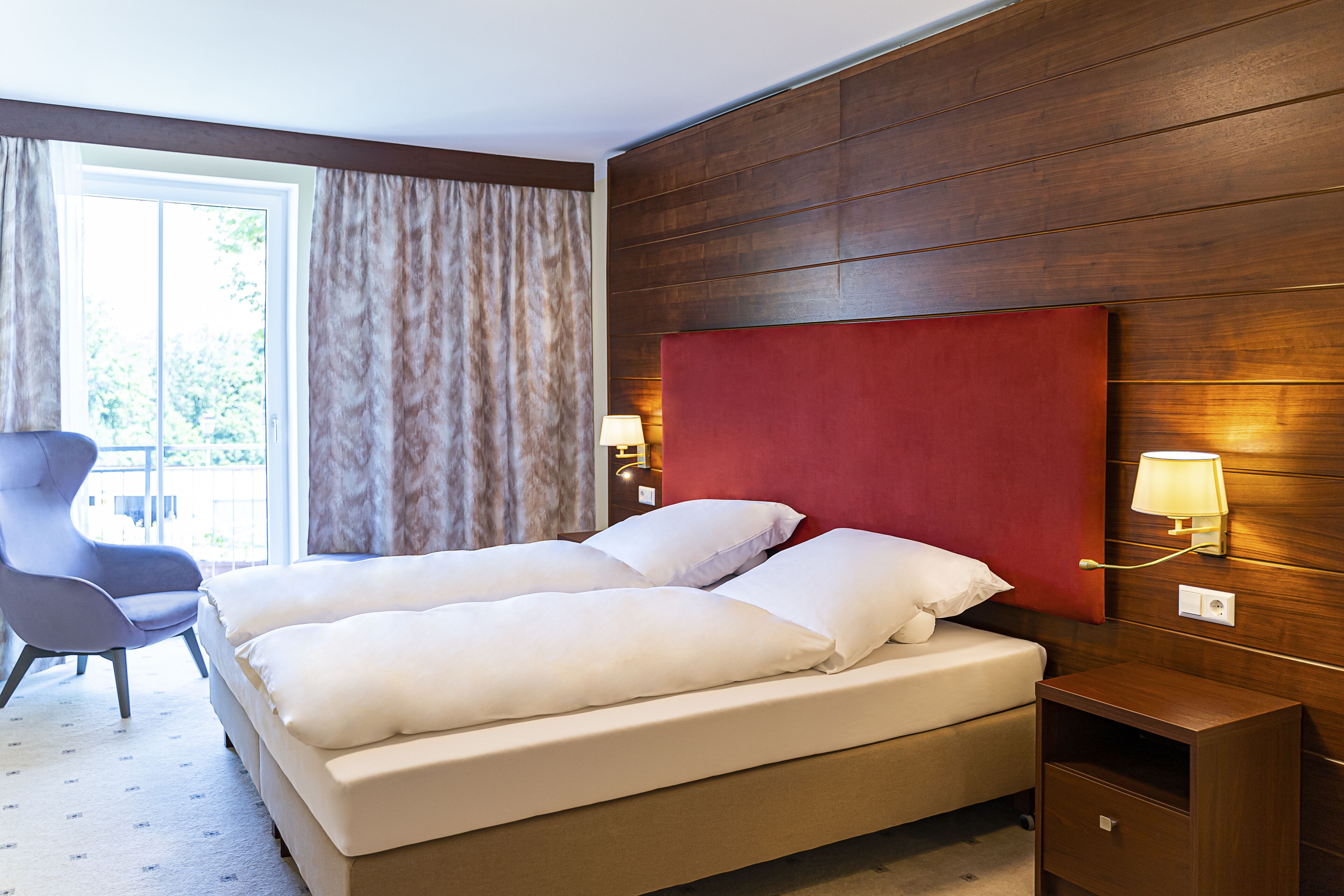 Une chambre d’hôtel avec un grand lit, une chaise, une table de chevet avec une lampe et une vue sur l’extérieur par une fenêtre.