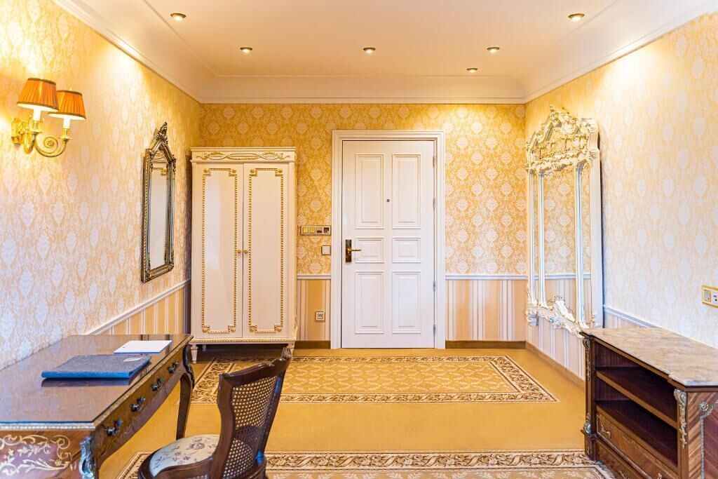 Una habitación con papel tapiz estampado, una puerta blanca, un escritorio con cajones, una silla y una pequeña mesa.