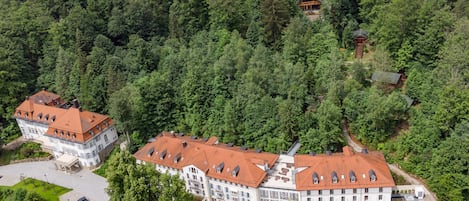 Un edificio grande de varios pisos con techo de tejas rojas, rodeado de un bosque denso.