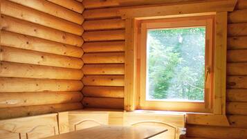 A wooden cabin interior with a dining table and benches, a window with a view of greenery, and a sloped ceiling.
