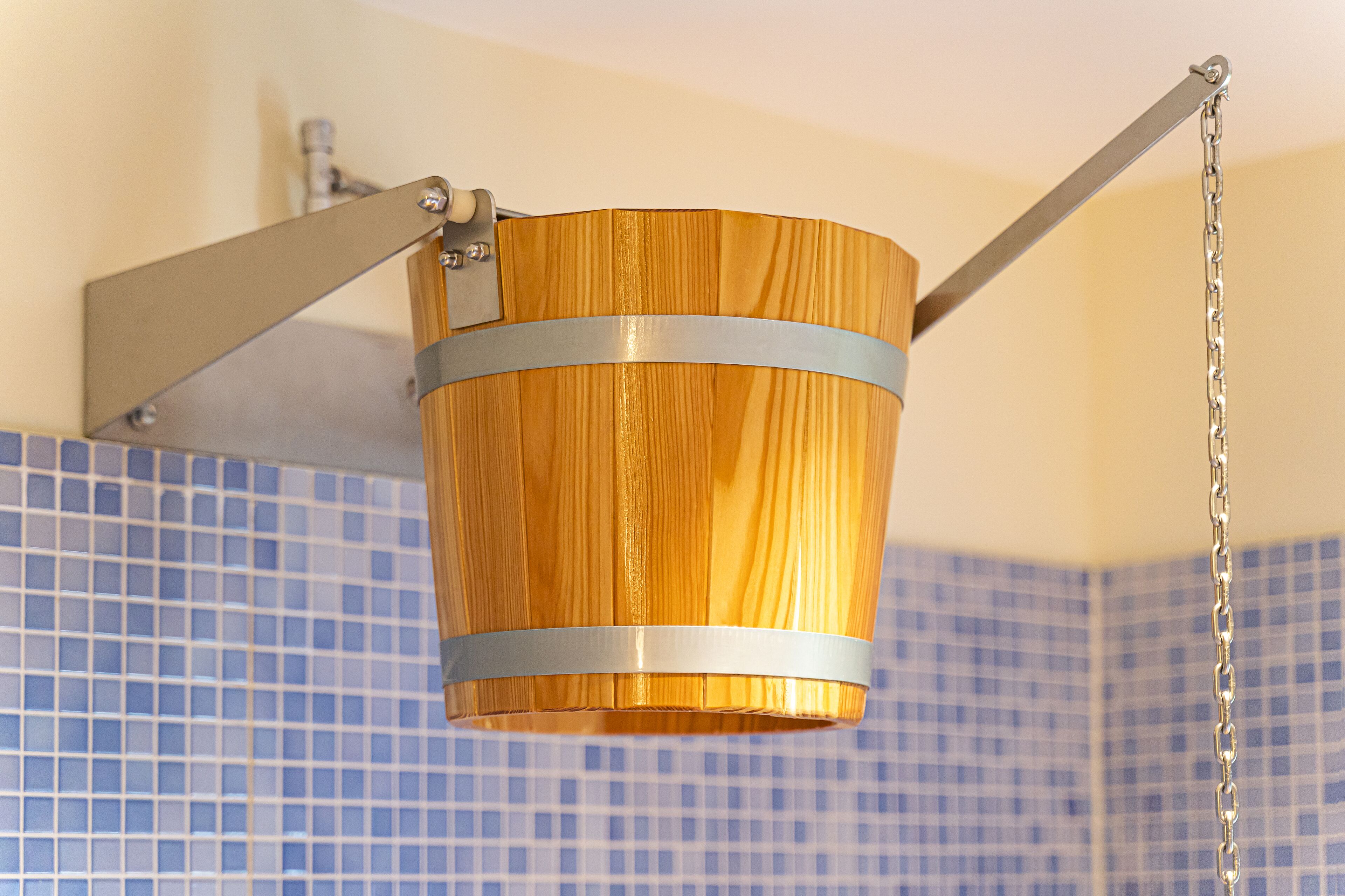 A wooden bucket suspended by a metal chain in a bathroom with blue tiled walls.