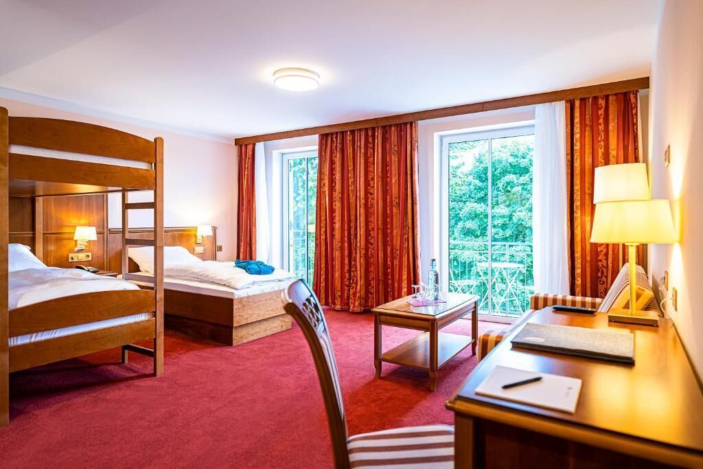 Habitación de hotel con dos literas de madera, un escritorio pequeño con silla, una mesa y vistas a zonas verdes a través de amplias ventanas.