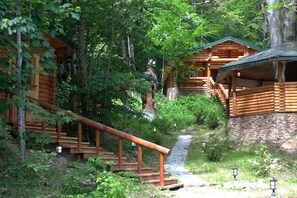 Una cabaña de troncos con cimientos de piedra, escaleras de madera y un pequeño arroyo.