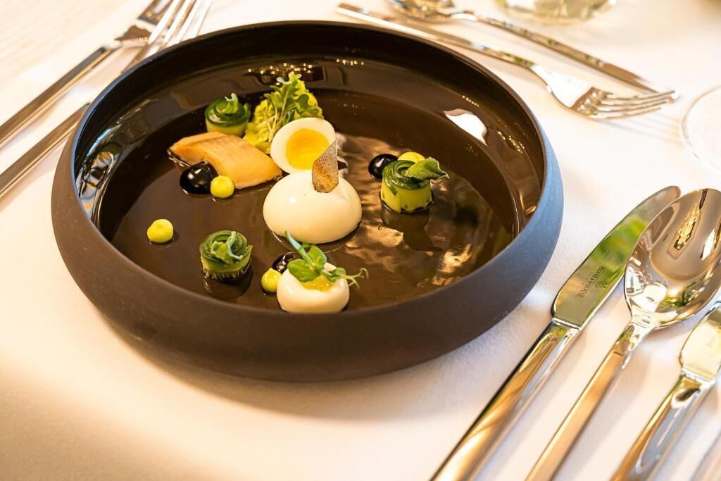 Un plato negro con un huevo blanco, hojas verdes y pequeños elementos amarillos sobre un mantel blanco.