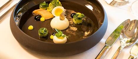 Un plato negro con un huevo blanco, hojas verdes y pequeños elementos amarillos sobre un mantel blanco.