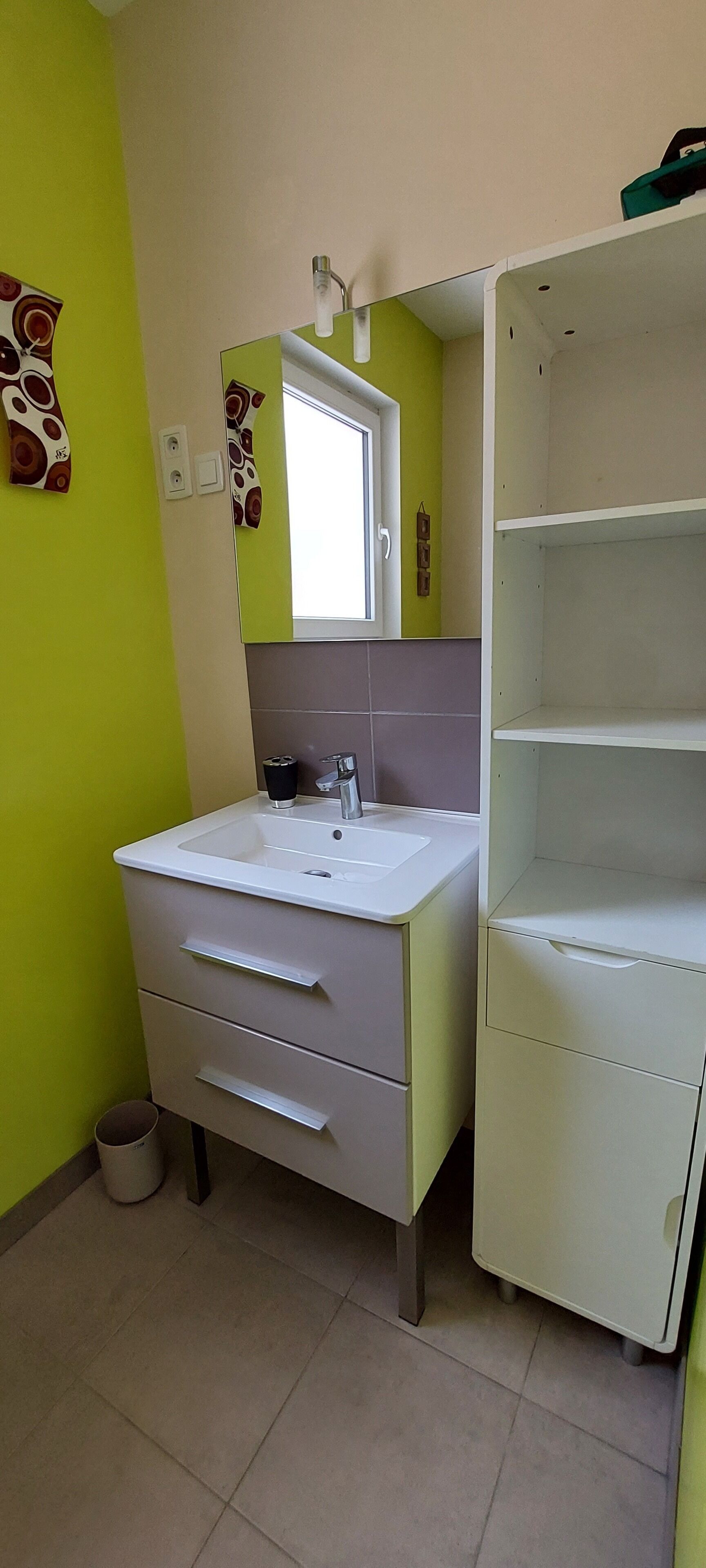Une salle de bain avec un lavabo blanc, un miroir et un mur vert. On y trouve un meuble blanc avec des tiroirs et une étagère.