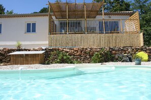 Ein Poolbereich mit Holzterrasse, Steinmauer und einem Gebäude mit Fenstern und einem überdachten Bereich.