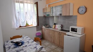 Une cuisine compacte avec des meubles en bois, un four à micro-ondes et un réfrigérateur. On y trouve une table à manger recouverte d’une nappe à motifs et une bouteille de vin. Une fenêtre avec des rideaux légers laisse entrer la lumière naturelle dans la pièce.