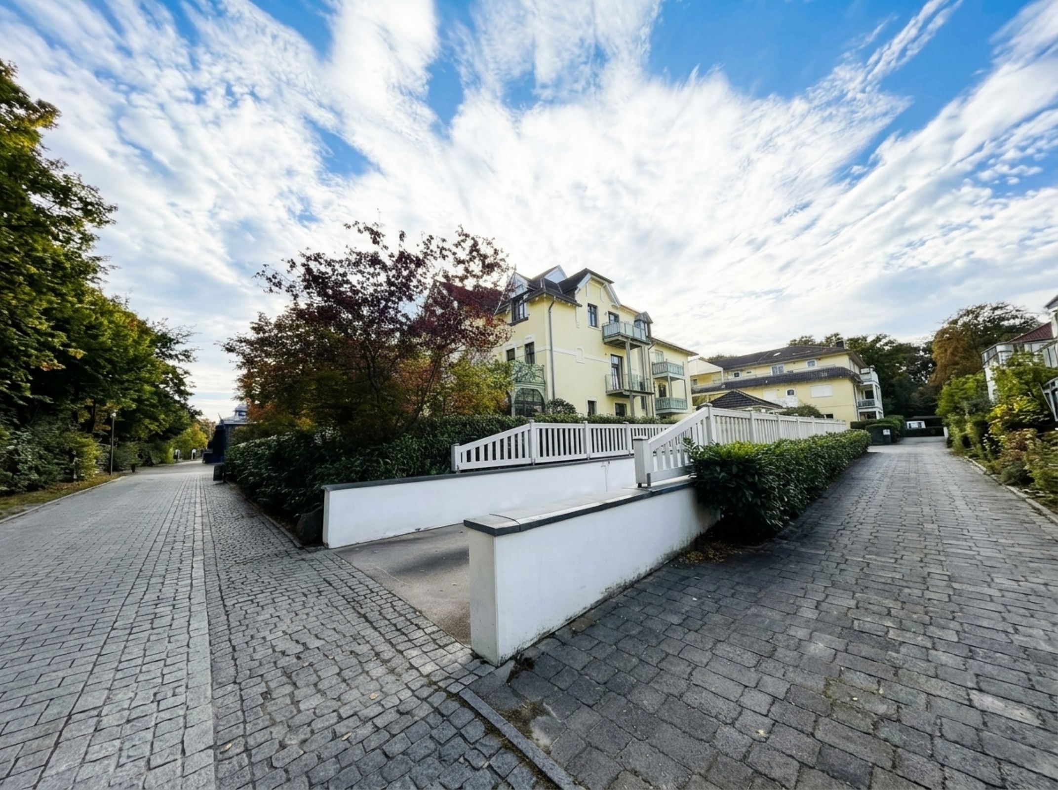 A residential street with a two-story house featuring a balcony, paved with cobblestones, and surrounded by greenery.