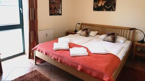 A bedroom with a bed, pillows, a red blanket, a radiator, a nightstand, a lamp, and a window with curtains. - Villa Laura 12 - Laura 12 (Kühlungsborn)