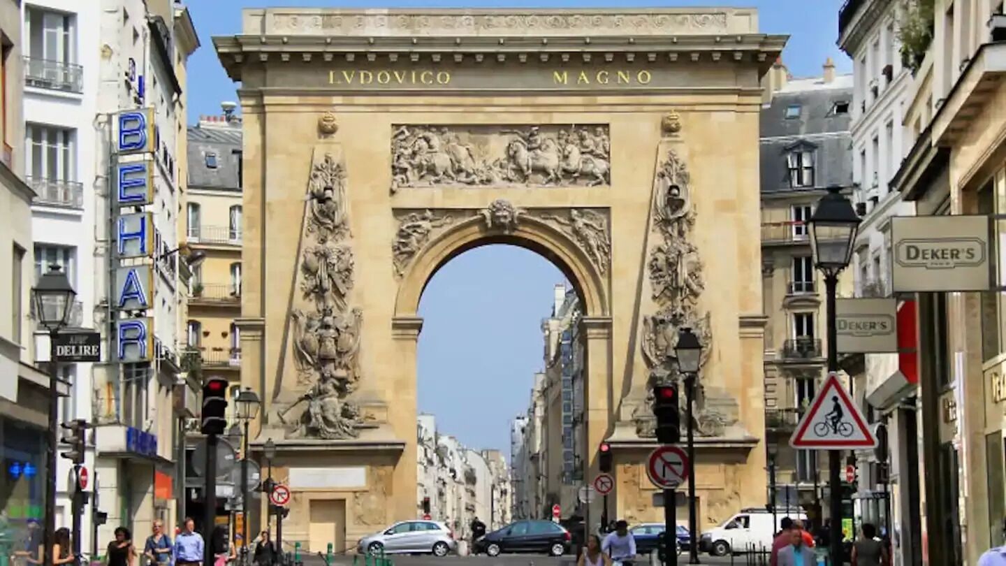 Un arco histórico con las inscripciones "LVDOVICO MAGNO" y "BEHRE" en sus laterales, rodeado de edificios urbanos y señales de calle.
