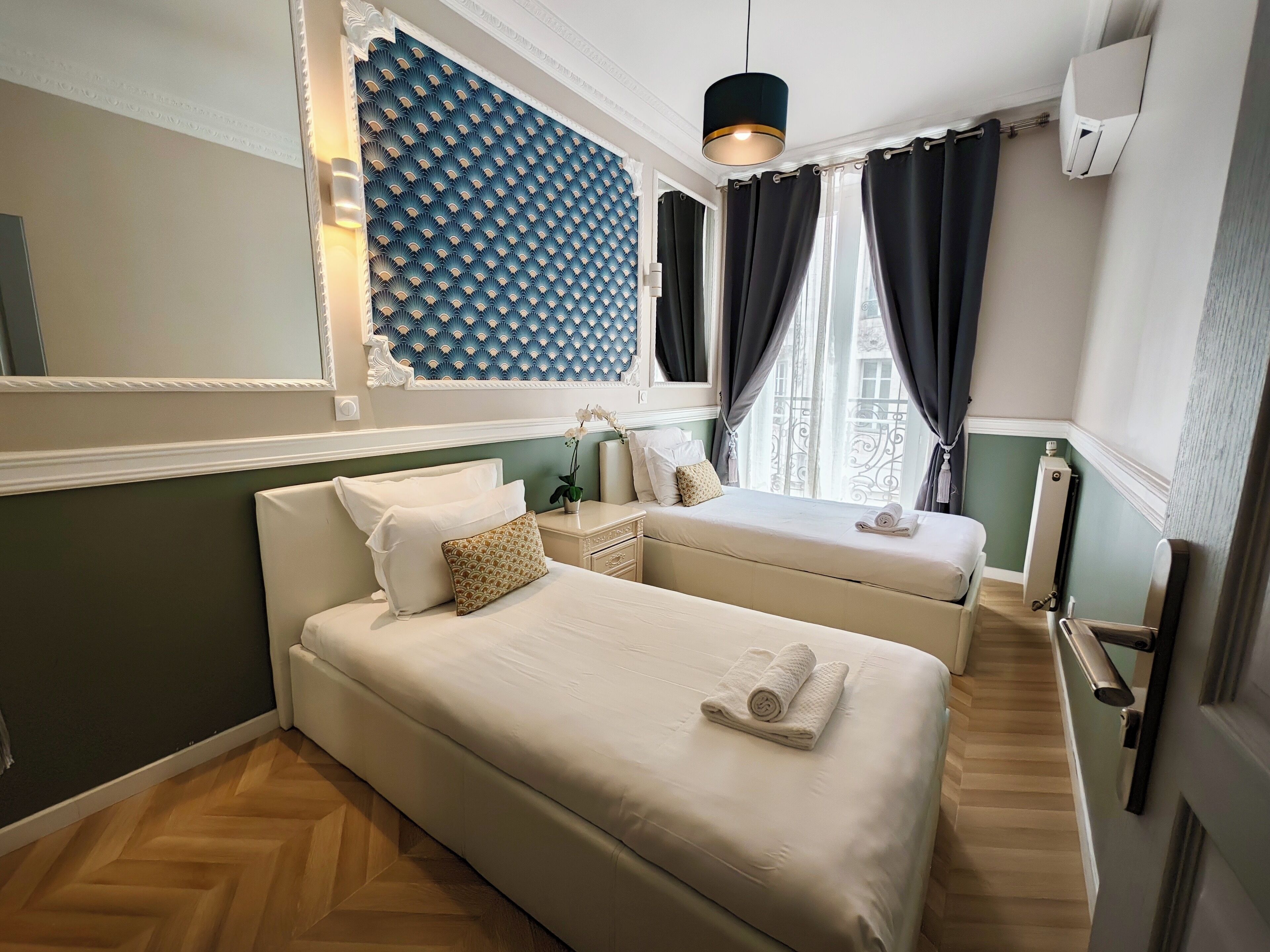 Een hotelkamer met een groot bed, een kleiner bed, een nachtkastje, een spiegel, een wandairconditioner en een raam met gordijnen.