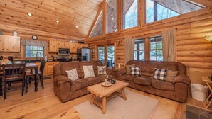L'intérieur de cette vaste cabane en rondins comprend un coin repas, deux canapés, une table basse en bois et de grandes fenêtres offrant une vue sur l'extérieur.