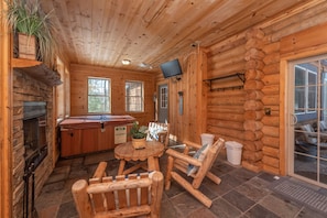 A log cabin interior with a stone fireplace, wooden rocking chairs, a hot tub, and a television mounted on the wall.