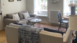 Un salón con sofá gris, sillón a rayas y una pequeña mesa redonda. Se ve una ventana con vistas a follaje exterior.