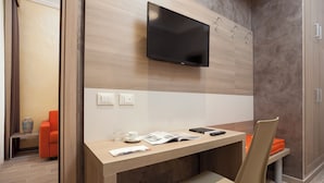 A modern hotel room with a wooden desk, a flat-screen TV mounted on the wall, and a seating area with a cushioned chair and orange cushion.