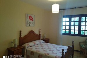 A bedroom with a wooden bed, a nightstand, a lamp, a window with curtains, and a wall-mounted artwork.