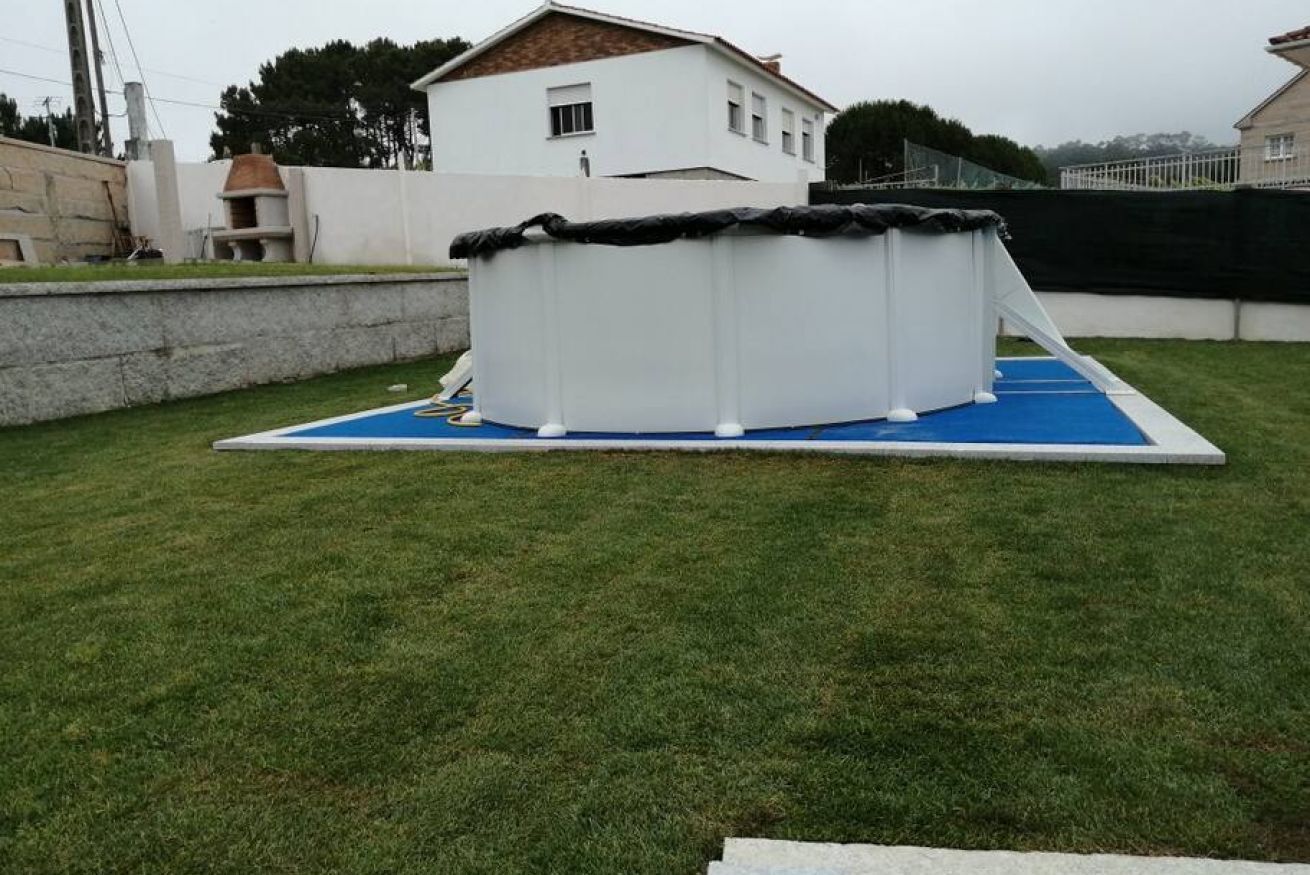 Ein Swimmingpool mit blauer Veranda und einer weißen Struktur darüber, umgeben von einer Rasenfläche und einem weißen Gebäude im Hintergrund.