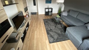 Una sala de estar moderna con un sofá gris, piso de madera, un televisor de pantalla plana, una mesa de centro y una puerta que da a otra habitación.