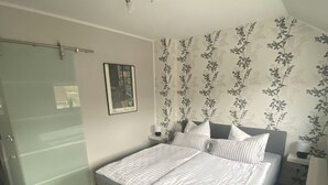 Un dormitorio con cama, mesita de noche, alfombra negra, pared con estampado floral y lámpara en el techo.