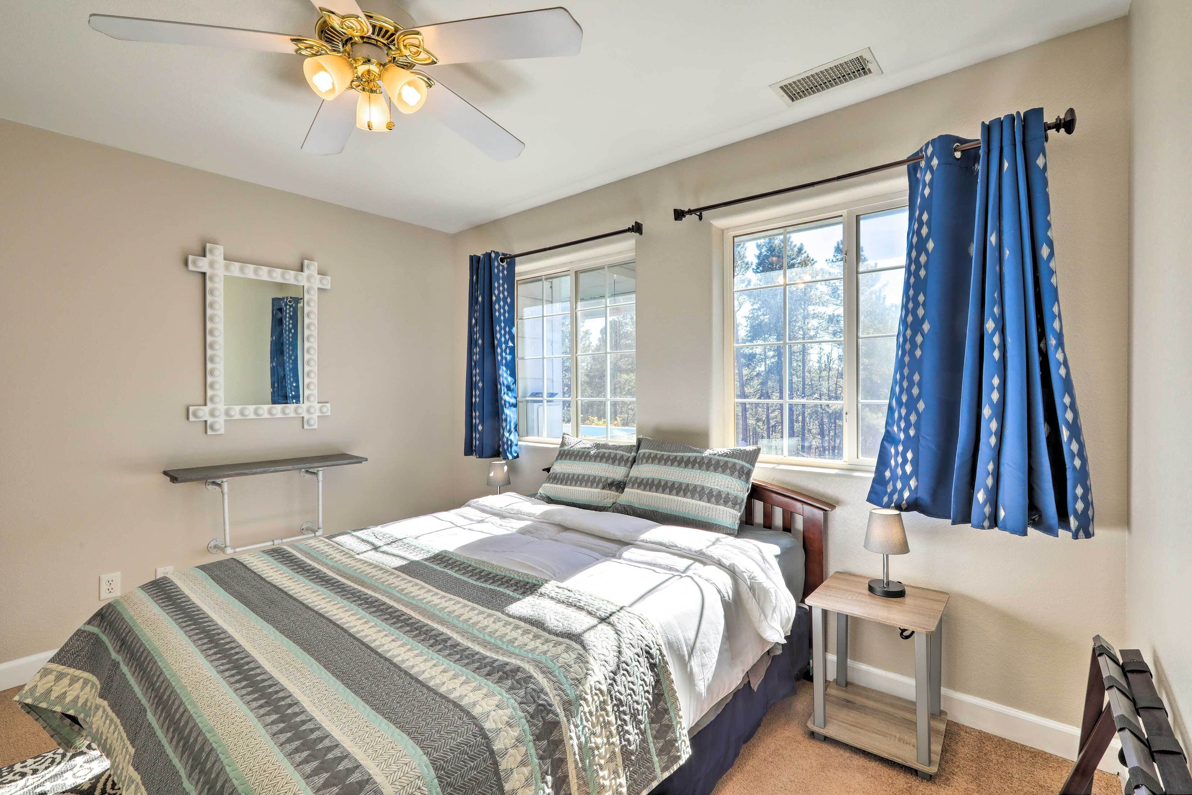 Ein Schlafzimmer mit Bett, Nachttisch mit Lampe, Deckenventilator, Fenster mit Vorhängen und Spiegel mit Beleuchtung.