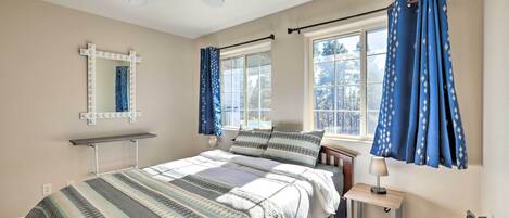 Ein Schlafzimmer mit Bett, Nachttisch mit Lampe, Deckenventilator, Fenster mit Vorhängen und Spiegel mit Beleuchtung.
