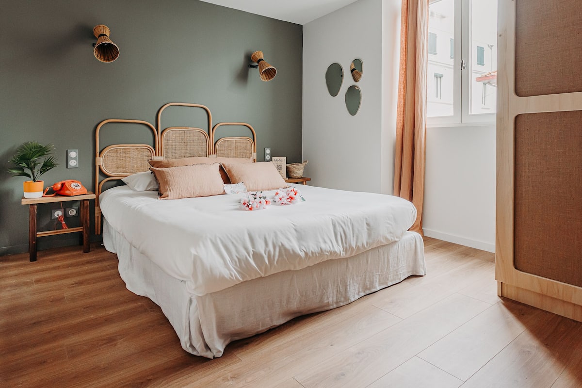 Un dormitorio con una cama grande, una mesita de noche con lámpara, una planta en maceta y un espejo montado en la pared.