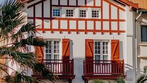A traditional building with a white facade and red timber framing, featuring balconies with wooden shutters and a red railing. - Palmito (Biarritz)