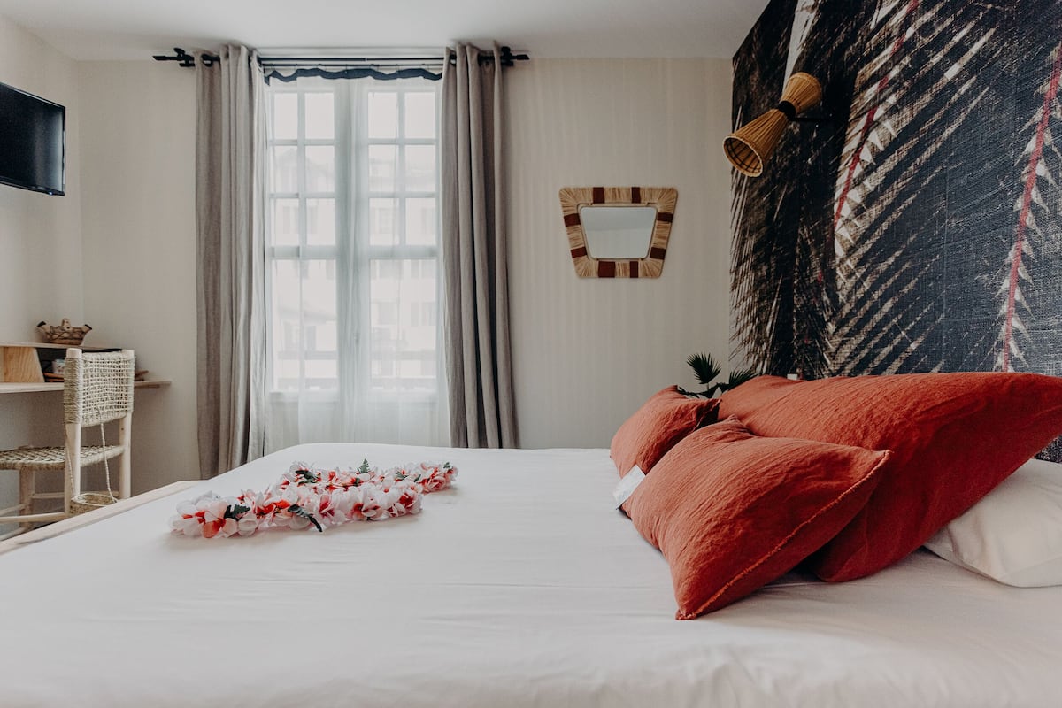 Una cama bien hecha con almohadas rojas y blancas, un arreglo floral y un espejo en la pared.
