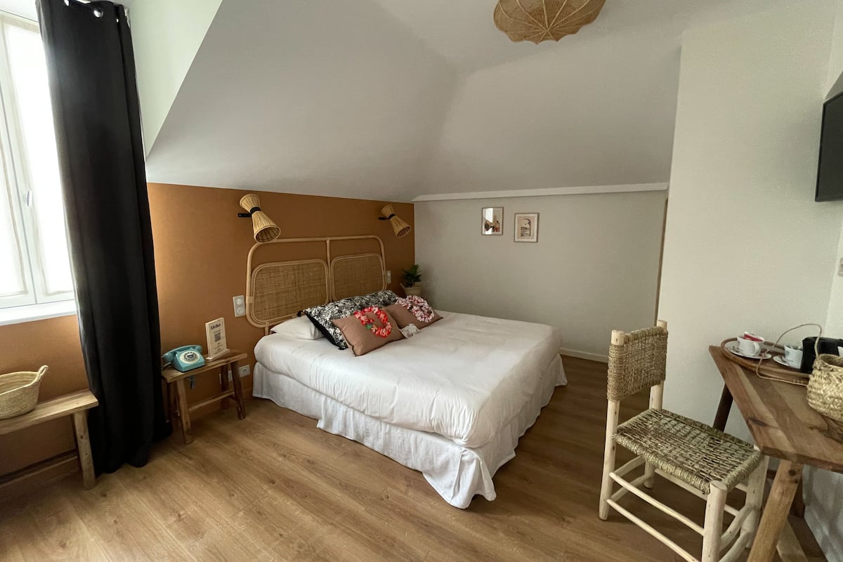 Un dormitorio con cama, mesitas de noche, una silla, una pequeña mesa y una ventana con cortinas.