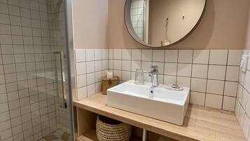 Een moderne badkamer met een douchecabine van glas, een witte wastafel met een ronde spiegel erboven en een houten opbergmeubel met een handdoekhouder.