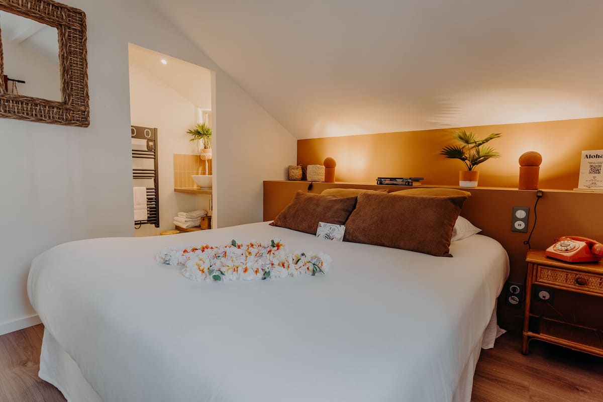 Una cama bien hecha con un arreglo floral, una mesita de noche con una lámpara y un espejo montado en la pared.