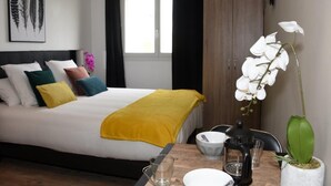 Une chambre à coucher bien rangée, avec un lit, des oreillers, une table de chevet sur laquelle se trouvent une cafetière, un bol et un verre.