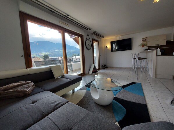 Un salón moderno con sofá, una mesa de centro de vidrio y vistas a montañas a través de amplios ventanales.