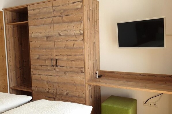 A bedroom with a wooden wardrobe, a bed, a green ottoman, and a mounted TV.