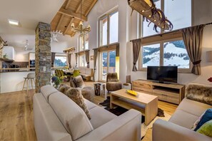 A spacious living area with a large sofa, a wooden coffee table, and a television. There is a stone pillar and a view of mountains through large windows.