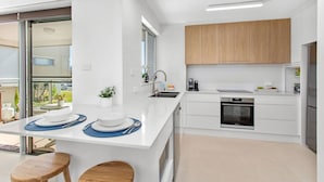 A modern kitchen with white cabinetry, a built-in oven, and a large island with a sink and a coffee maker. - Waves on North - El Sandi 4 (Forster)