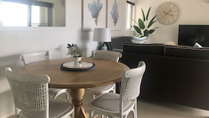 A modern living room with a round wooden dining table surrounded by white wicker chairs, a dark sofa, a large mirror, and a wall clock. - Waves on North - El Sandi 4 (Forster)