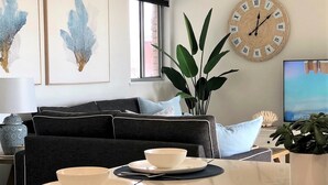 A living room with a dark sofa, a dining table set with bowls and plates, a large wall clock, and a plant. - Waves on North - El Sandi 4 (Forster)
