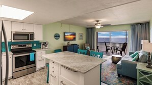 Cocina compacta con microondas, horno y una pequeña isla. Hay una zona de comedor con sillas y mesa, y un balcón con vistas.
